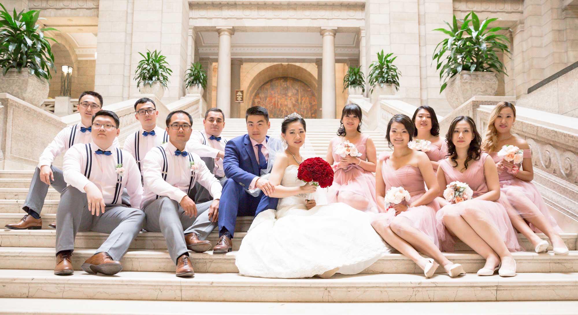 Image description: A group photo of a wedding of 12 people, with 5 groomsmen, 5 bridesmaids and the bride and groom. On the stairs of the Manitoba Legislative Building in Winnipeg.