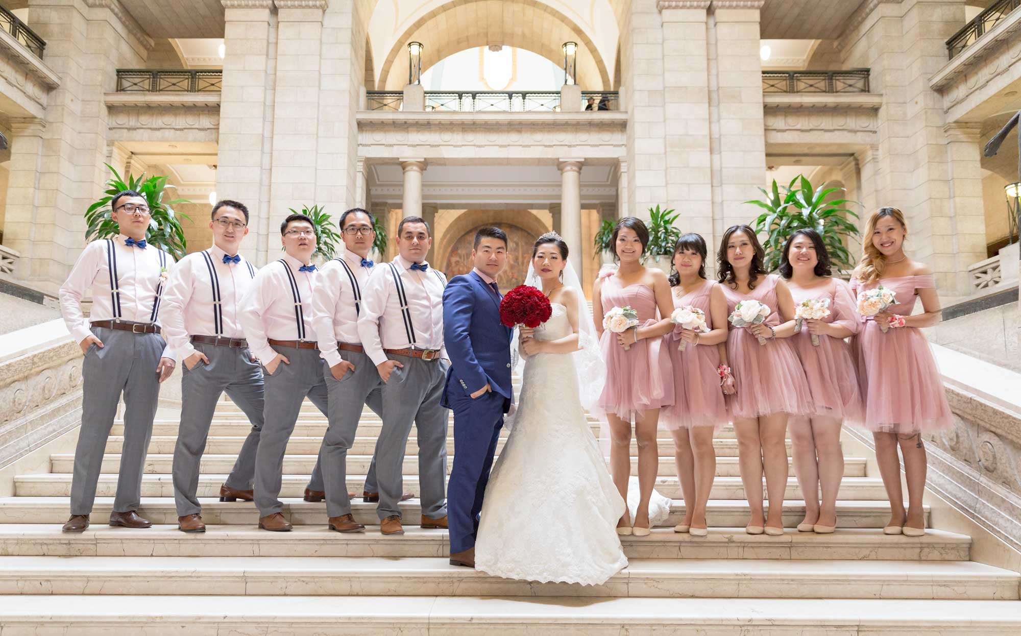 Image description: A group photo of a wedding of 12 people, with 5 groomsmen, 5 bridesmaids and the bride and groom. On the stairs of the Manitoba Legislative Building in Winnipeg.