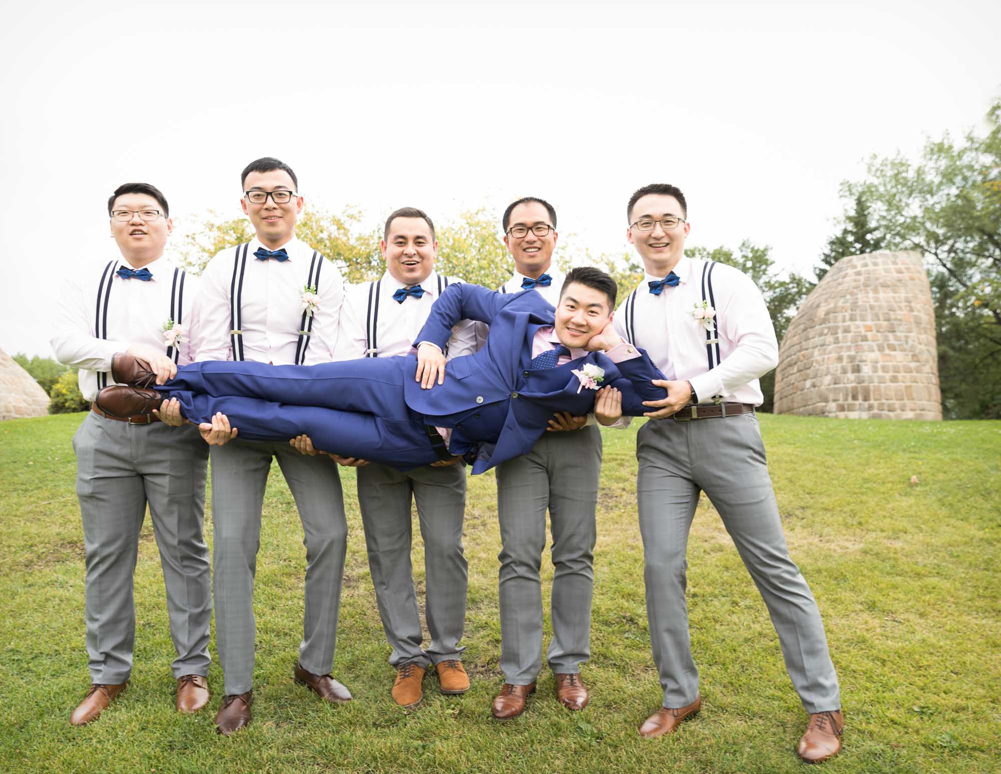 Image description: Five groomsmen in white shirts and grey trousers lifted the groom in a blue suit on the green grass.