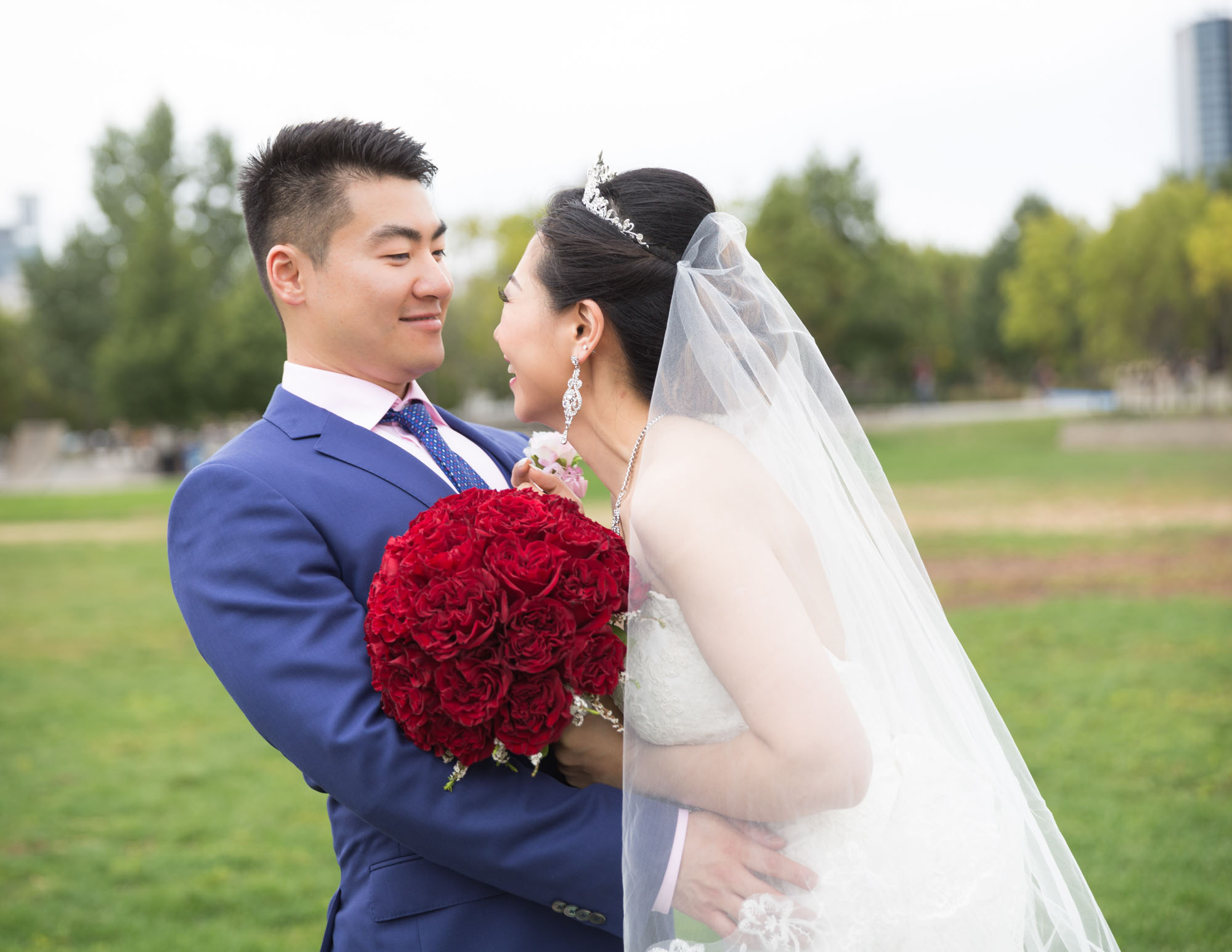 Image description: A photo of the bride and groom, the bride smiling at the groom and holding a bouquet of red roses.