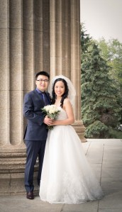 Image description: The groom in a blue suit and the bride in a white wedding gown carrying a white bouquet smiled for the camera.