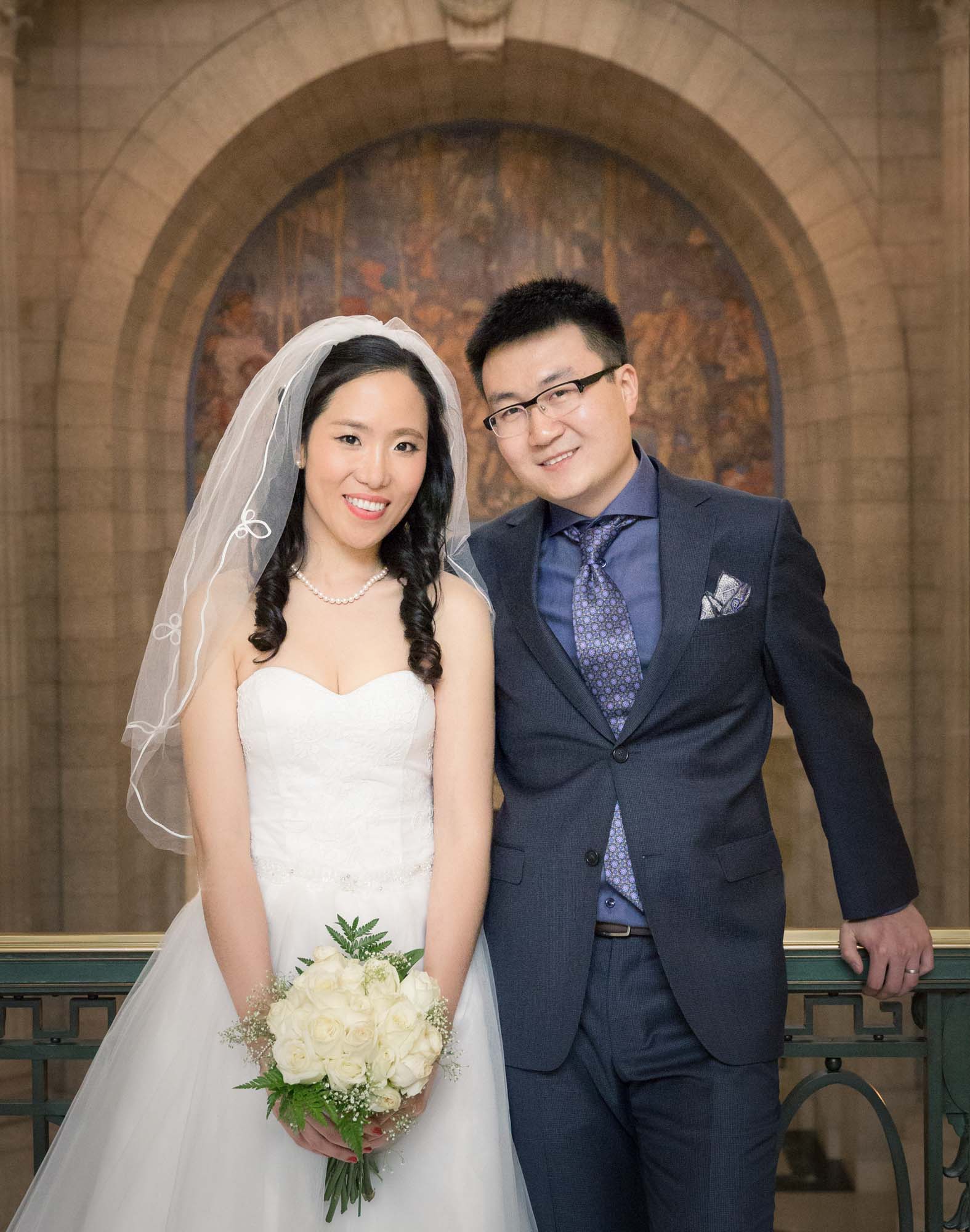 Image description: The groom in a blue suit and the bride in a white wedding gown carrying a white bouquet smiled for the camera.