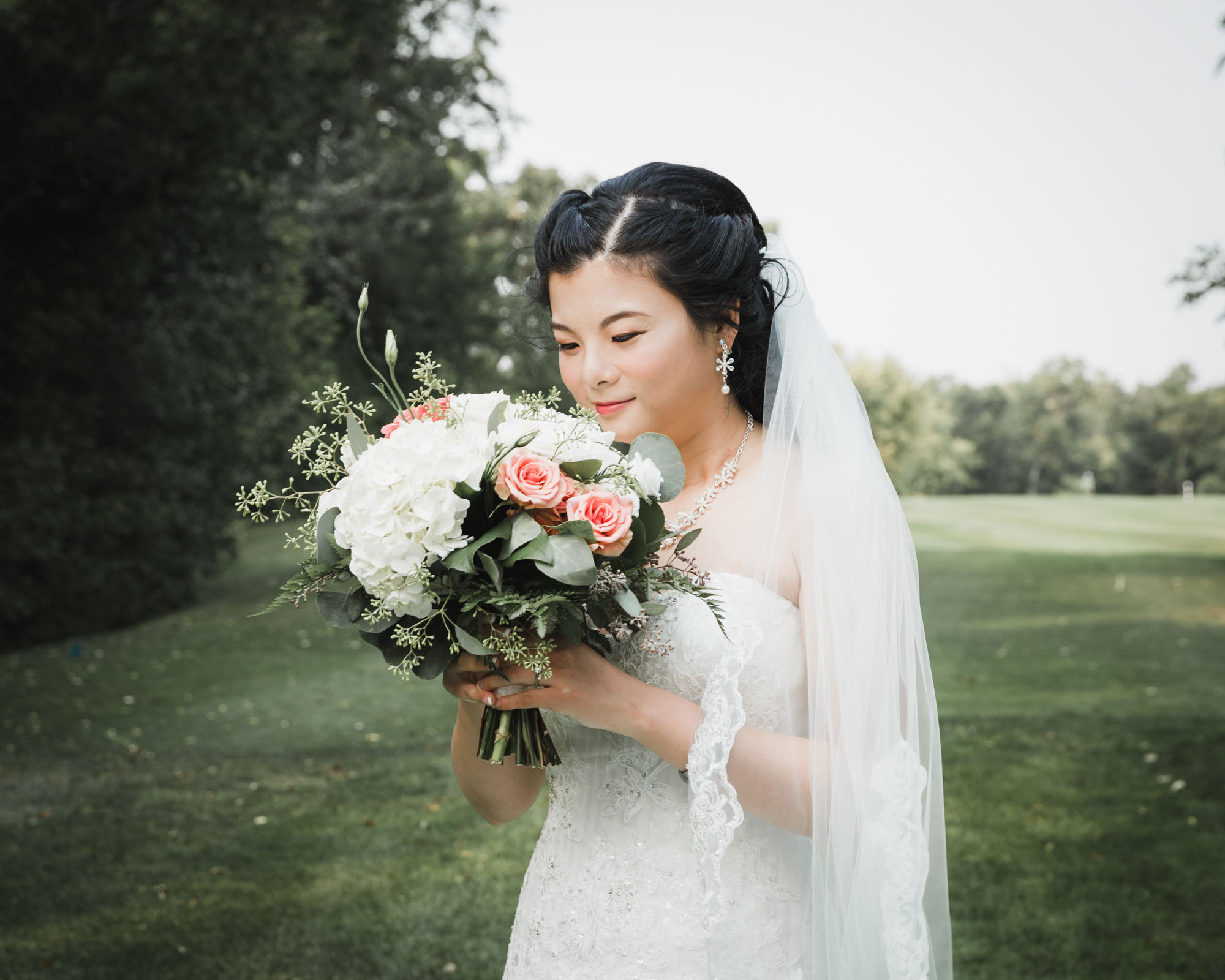 Image description: The bride looked at his bouquet and wore a white wedding dress on the green grass against the blue sky.