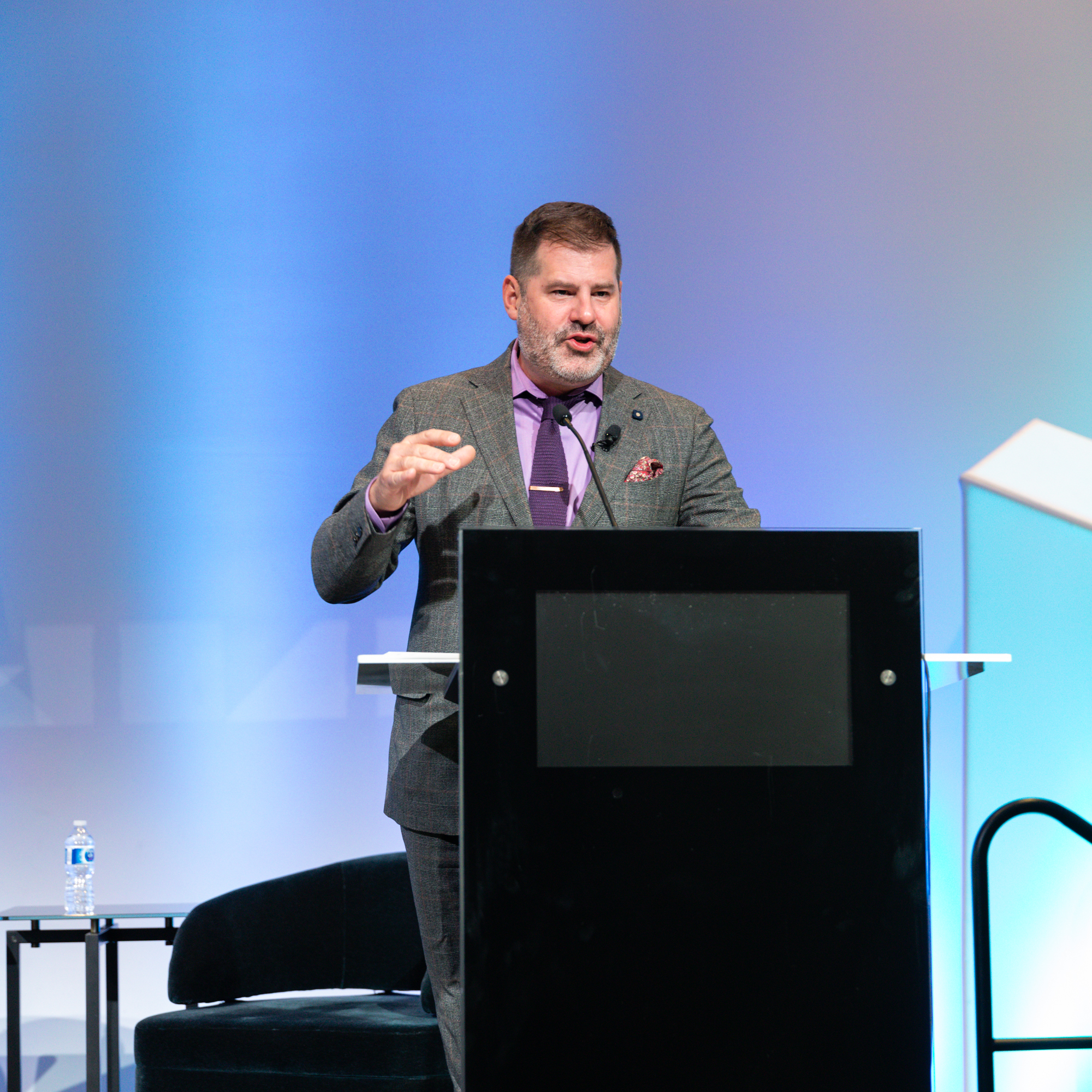 Image description: a man in a gray suit with a purple shirt is standing on a speech stage. Purple background.