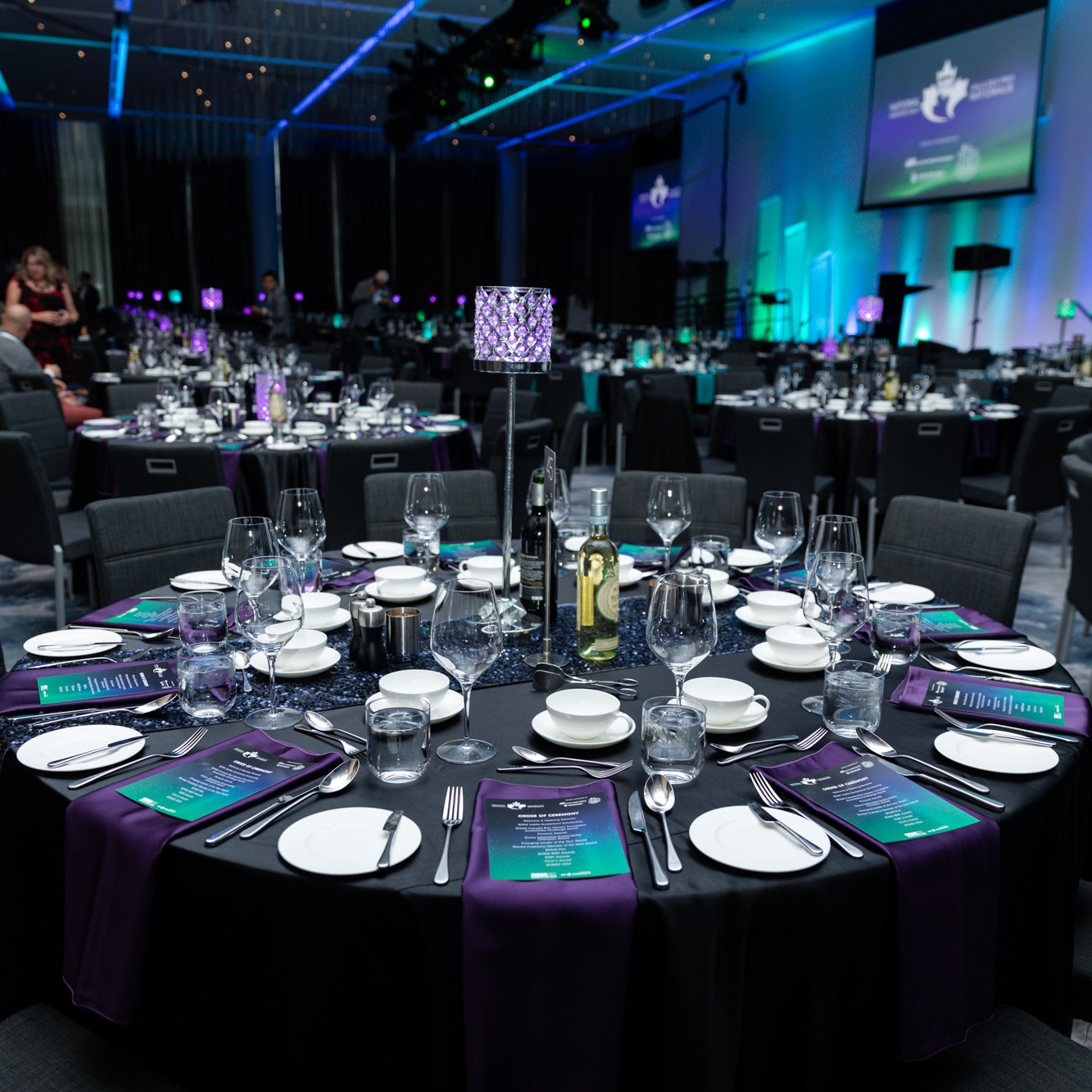 Image description: In a venue there are many round tables, each table is arranged with the schedule of events and drinks and champagne. Text reads: BOMEX2023