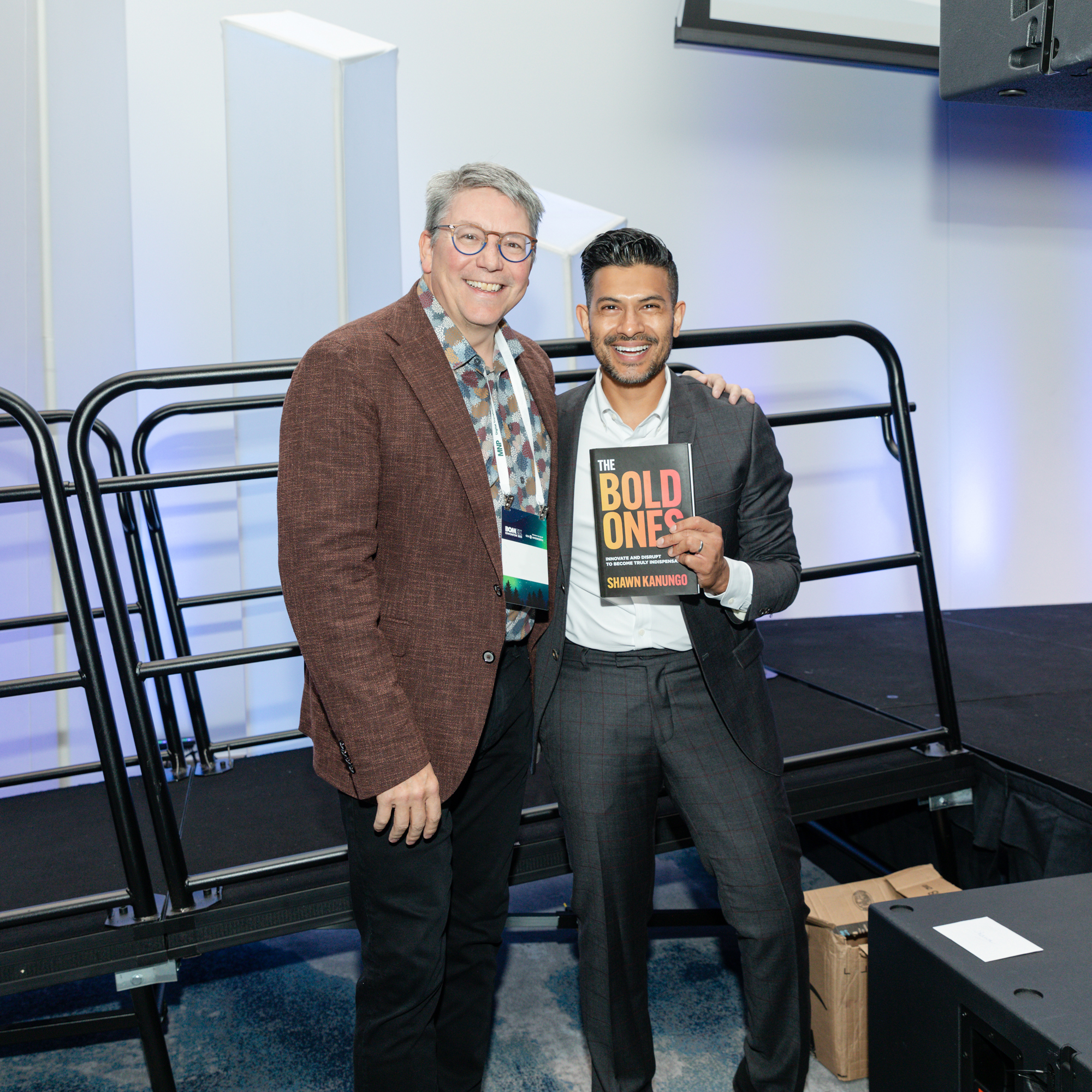 Image description: A photo of two men. One of them, a man in a black yellow and white shirt, was holding a book.Text reads: BOLD ONES By Shawn Kanungo