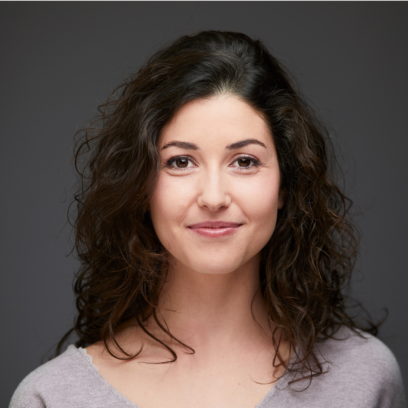 Image description: A headshot of a woman wearing a light gray top, curly hair. Dark grey background.