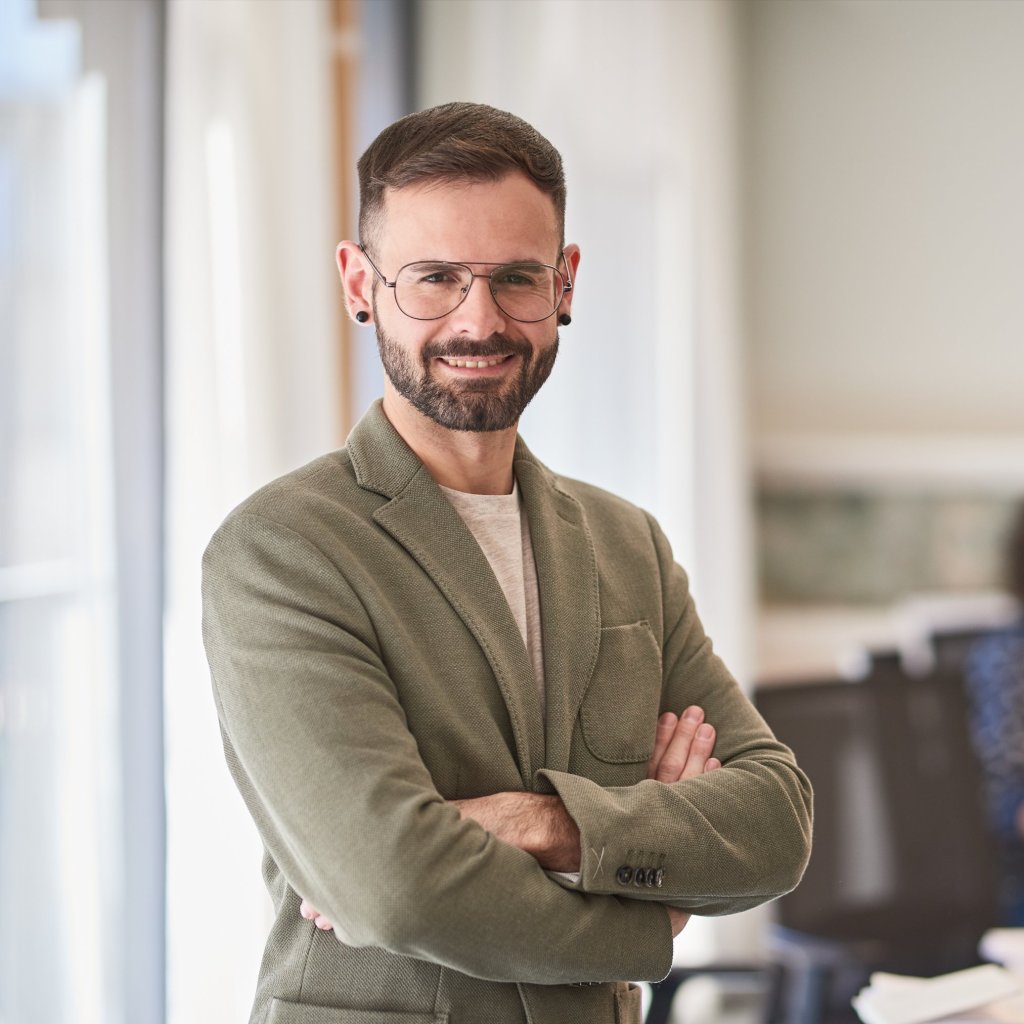 Image description: A headshot of a man wearing a light gray top, with glasses. An office setting background.